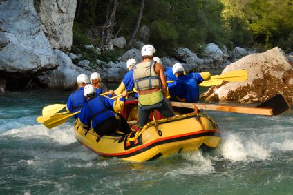 A group of people in an inflatable raft are navigating river rapids in a canyon. The leader stands in the aft of the raft, assessing the risks ahead and steering the raft through the hazards.